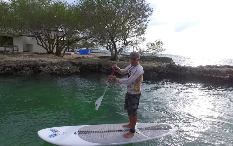 Remador con camiseta de Colombia Flyboard sobre un paddle board blanco, en aguas verde turquesa junto a una isla cerca de Cartagena